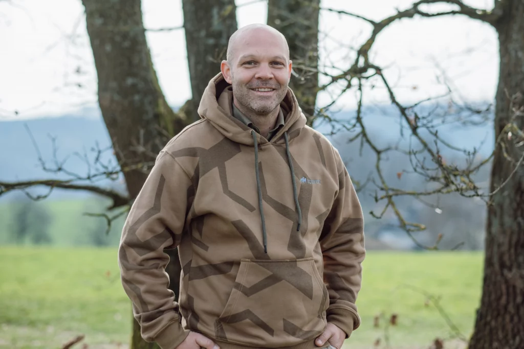 Nico Kessler-Thönes steht in einem braunen Hoodie vor einem Baum mit natürlichem Hintergrund und lächelt in die Kamera, Jagdausbilder der Hochsitz Akademie