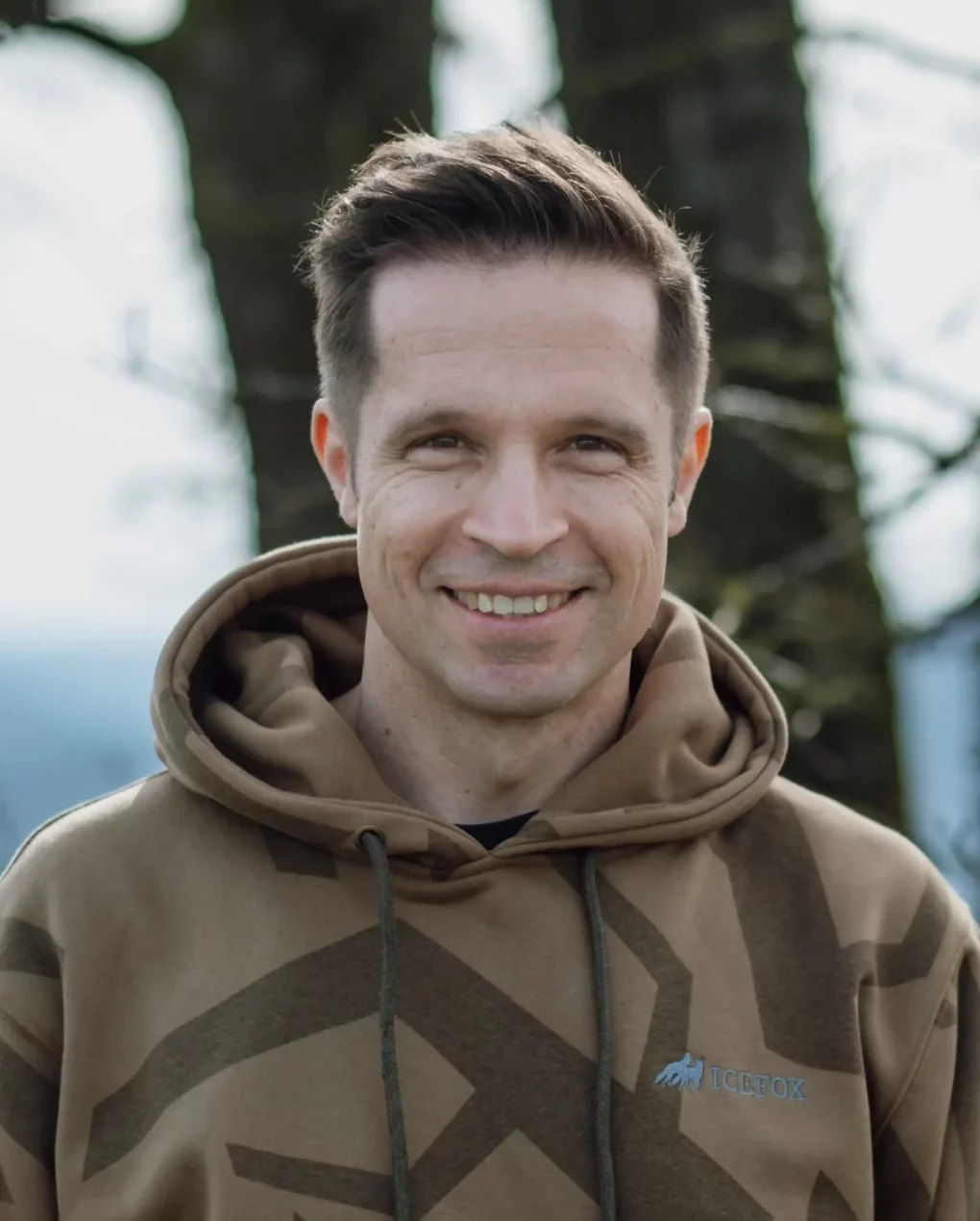 Dr. Tino Kessler-Thönes trägt einen braunen Hoodie und steht vor einem Baum mit unscharfem Hintergrund in der Natur, Jagdausbilder der Hochsitz Akademie