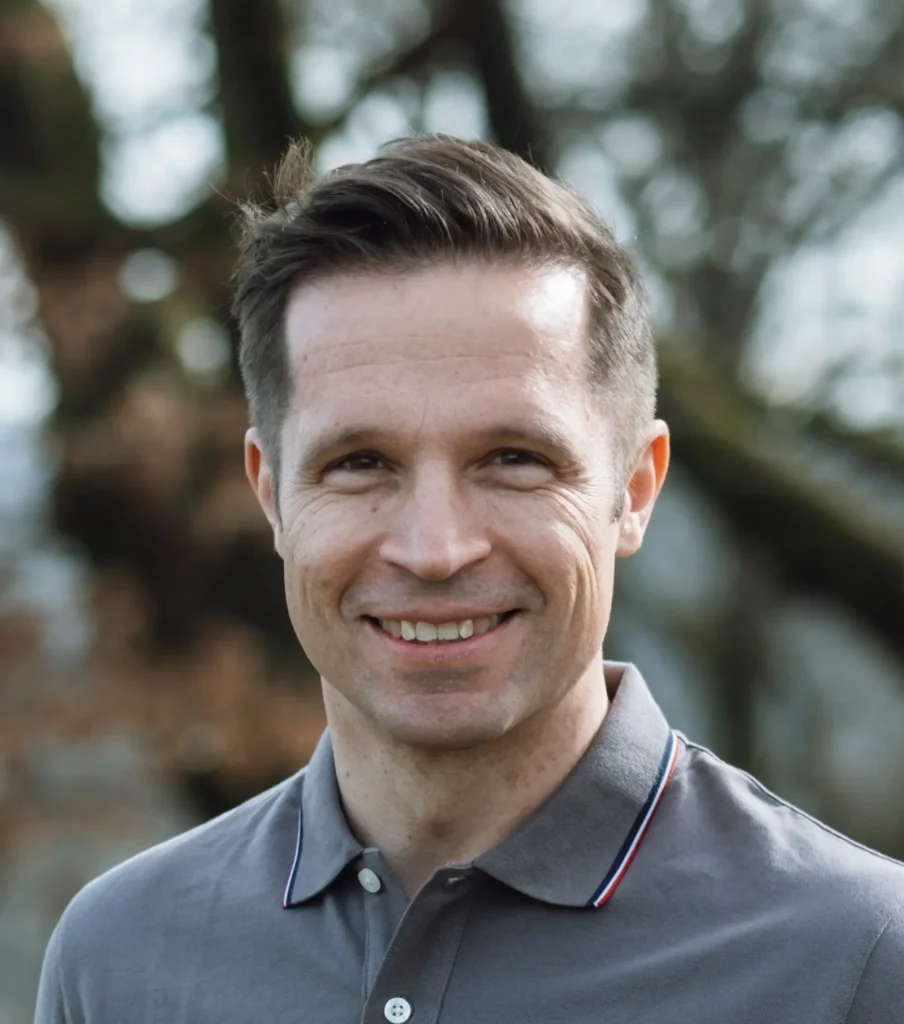 Dr. Tino Kessler-Thönes lächelt in die Kamera und trägt ein graues Polo-Shirt, unscharfer Hintergrund mit Bäumen, Jagdausbilder der Hochsitz Akademie