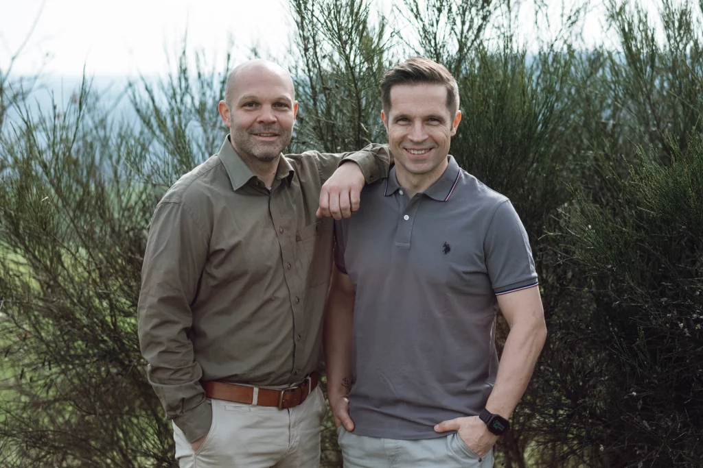 Dr. Tino und Nico Kessler-Thönes stehen eng nebeneinander vor Büschen in der Natur und lächeln in die Kamera, Jagdausbilder der Hochsitz Akademie